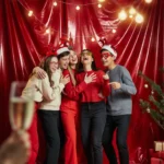 A group of teens laughing mid-photo in front of a shimmering red foil backdrop, wearing festive headbands and novelty glasses, string lights glowing in the background.