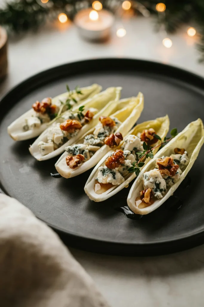 Small endive leaves filled with crumbled blue cheese, candied walnuts, and a drizzle of honey, fanned out on a dark plate. class=