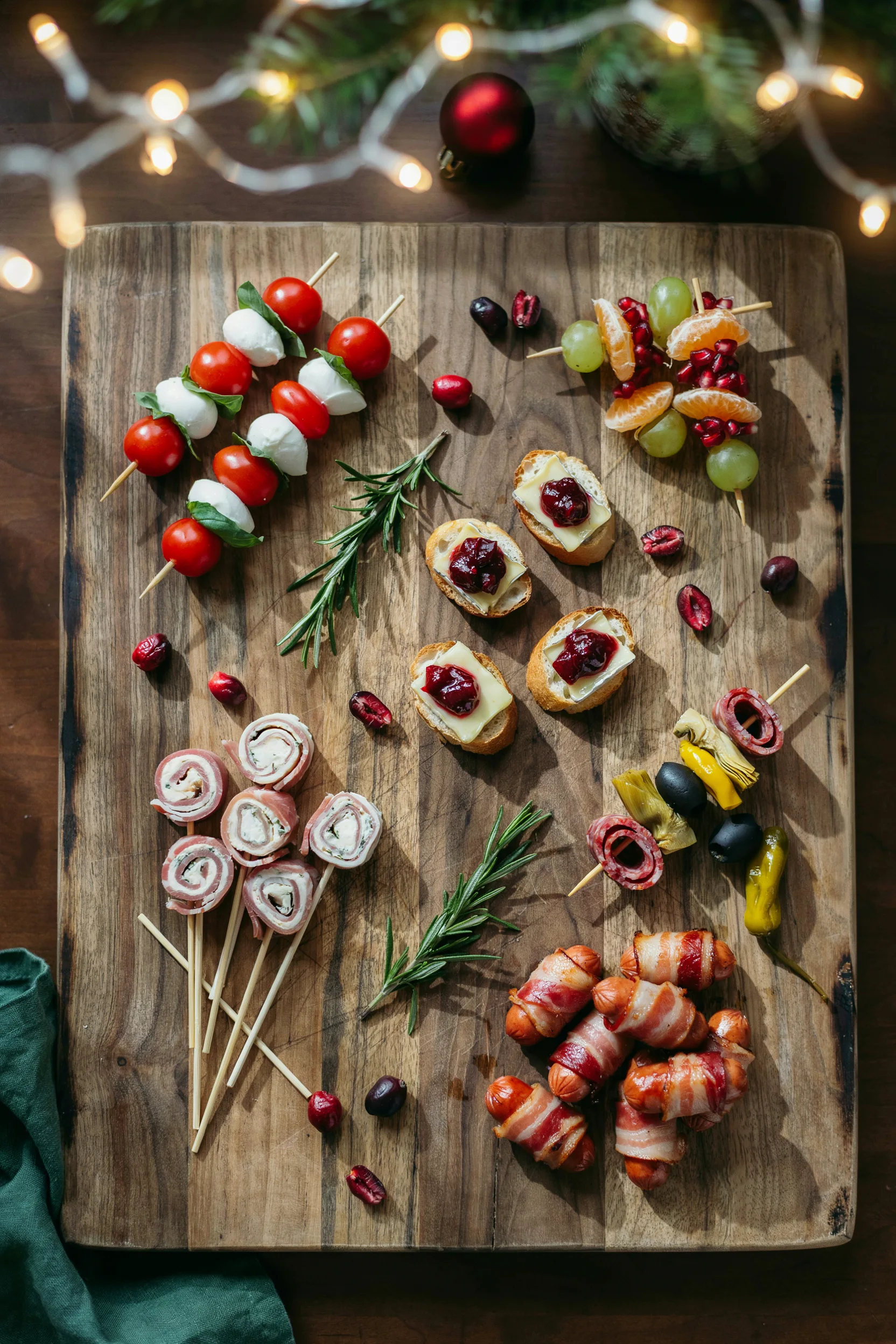 An overhead shot of a large wooden platter arranged with several types of toothpick appetizers — caprese skewers, cranberry brie bites, ham pinwheels — styled with sprigs of rosemary and a few loose cranberries scattered between them, warm string lights softly blurred in the background.]