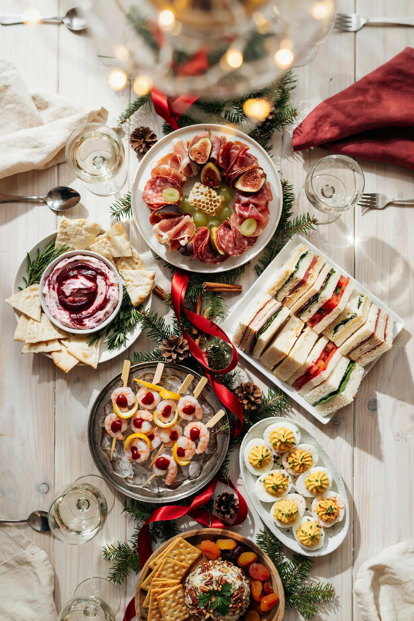 An overhead shot of a fully spread holiday buffet table showing several cold appetizers at once — a charcuterie wreath, a pink dip, pinwheel spirals, and skewers on ice — styled with pine sprigs and red ribbon, warm string-light glow.