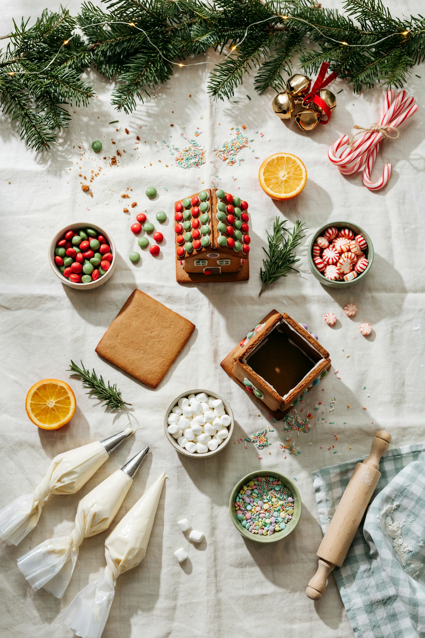 : An overhead shot of a kids' gingerbread decorating table mid-party — houses in progress, bowls of candy, small hands reaching for gumdrops, warm string lights in the background.