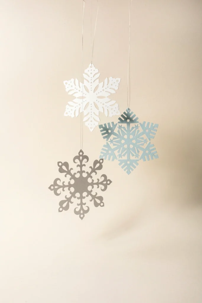 Three delicate paper snowflakes in white, pale blue, and silver hanging on clear fishing line with intricate cut patterns visible