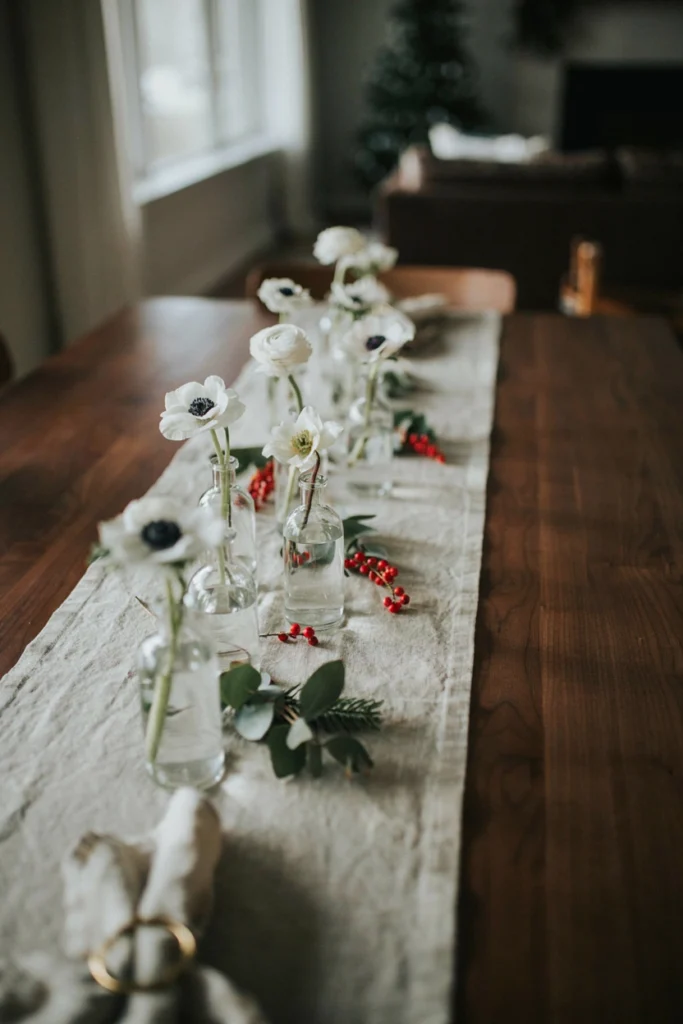 Row of small glass bud vases, each holding a single white flower stem, spaced along a plain linen runner