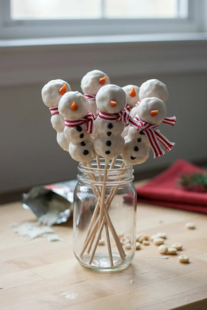Round rice cereal treat balls on sticks, coated in white candy melts with a scarf and stick arms, resembling snowmen.