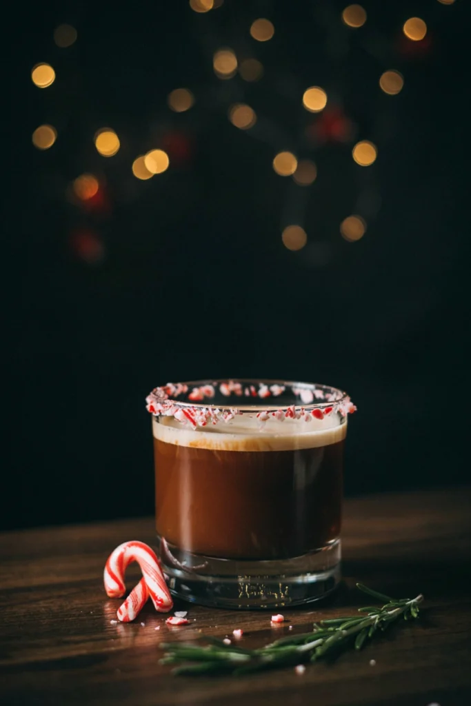 A rocks glass with layered cream and dark coffee liqueur, crushed candy cane pieces rimmed around the top edge, dark moody background
