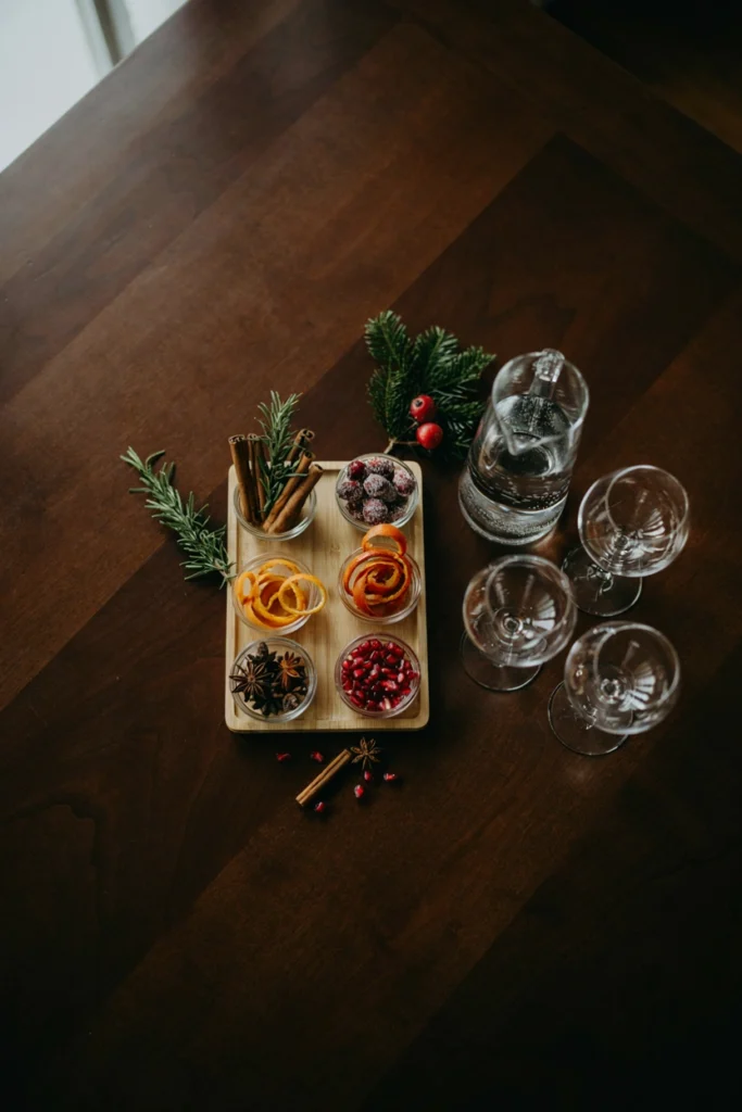 A wooden tray with garnishes including cinnamon sticks, rosemary, sugared cranberries, orange twists, and star anise in tiny glass bowls